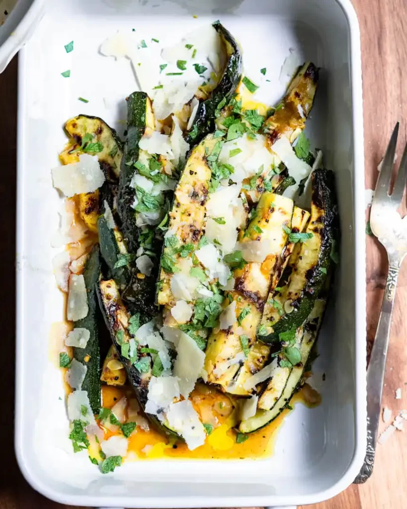 Grilled zucchini spears topped with parmesan cheese shavings and minced fresh parsley in a serving dish.