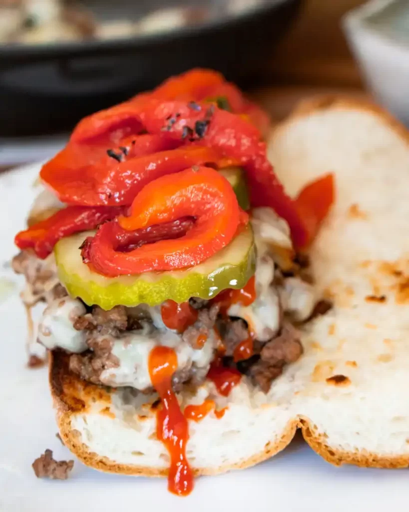 Open faced sandwich with ground beef, melted American cheese, pickles, peppers, and ketchup on top of it.