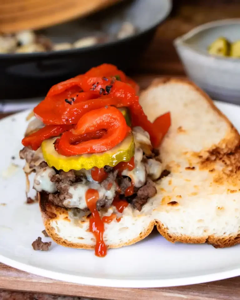 A Philly Cheese steak sandwich made with ground beef on a plate. The sandwich has cheese, pickles, ketchup and a roasted pepper topping.