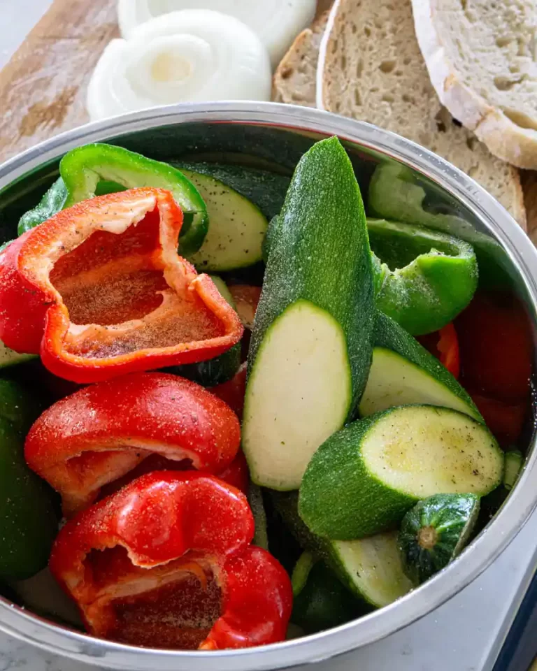 Raw red and green peppers cut in half with seeds removed. Along side the peppers are long diagonal slices of raw zucchini. All the veggies have a light seasoning on them.