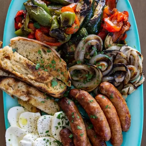 A platter filled with grilled veggies, grilled sausages, sliced mozzarella cheese, and sliced toasted bread.