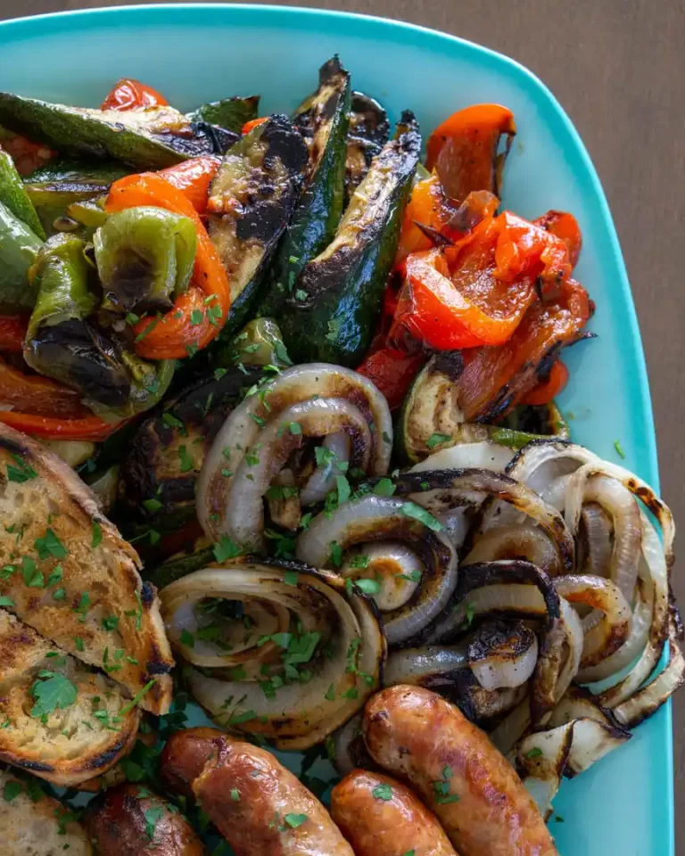 Grilled onions, peppers and zucchini on platter alongside Italian sausage.