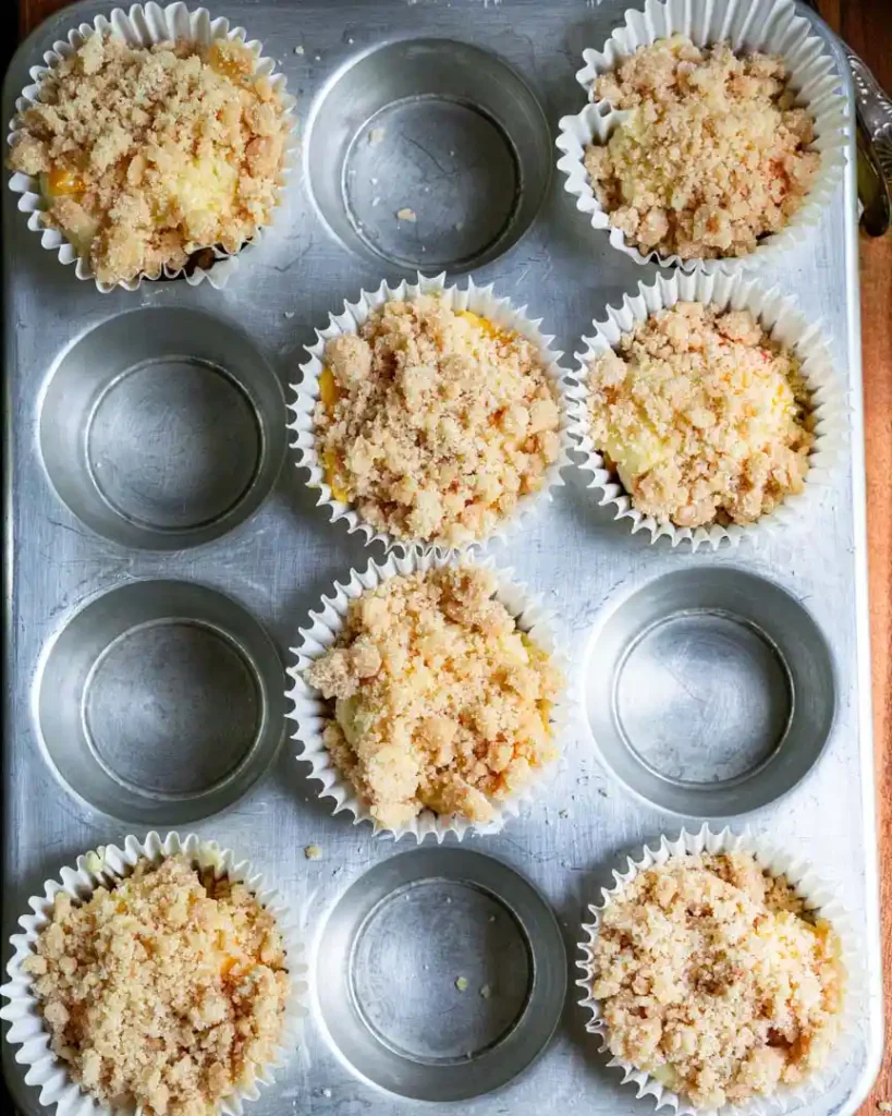 A muffin pan with unbaked peach muffins with streusel topping.