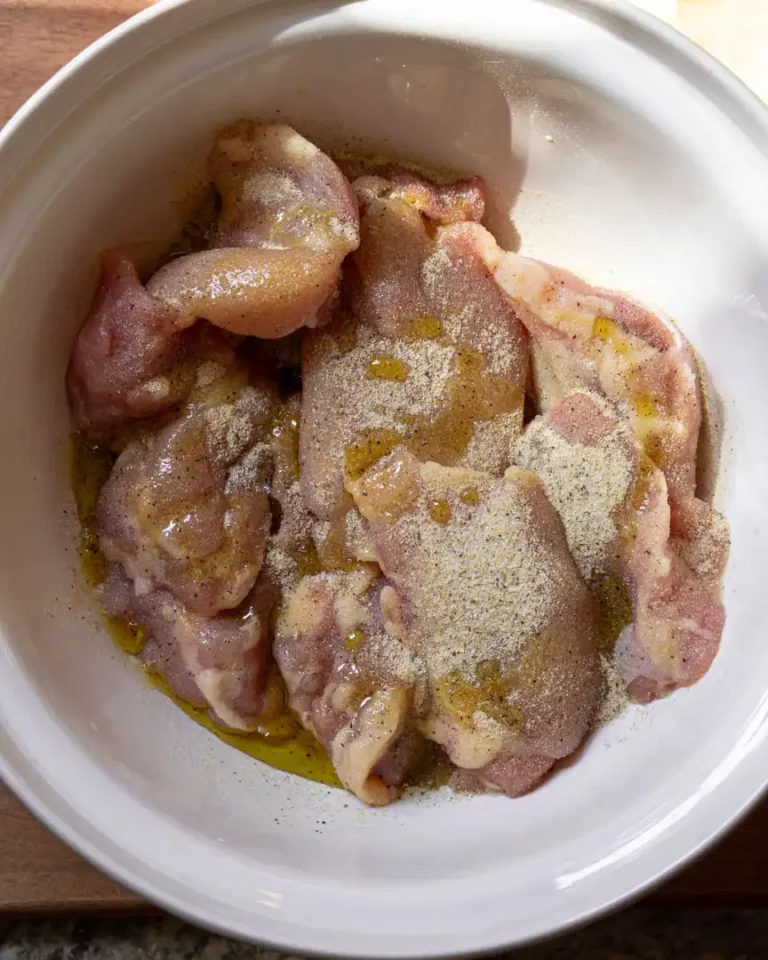 Uncooked chicken thighs in a bowl with seasoning and olive oil drizzled over them.