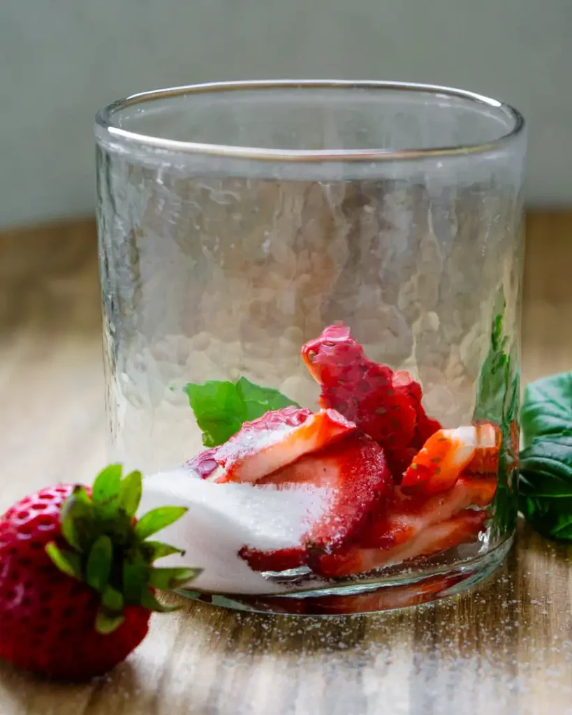 A short glass filled with sliced strawberries, fresh basil, and sugar. There is a whole strawberry and some basil leaves next to the outside of the glass.