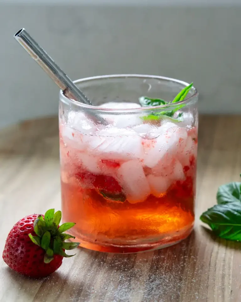 A short glass filled with strawberry basil spritzer mocktail. The glass has a stainless steel straw coming out of it. There is a strawberry and basil leaves next to the outside of the glass.