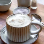 A mug of maple sea salt latte. The mug sits on a small plate. there are small pitchers and a mini bowl of salt behind the mug.