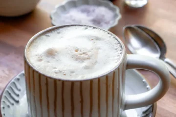 A mug of maple sea salt latte. The mug sits on a small plate. there are small pitchers and a mini bowl of salt behind the mug.