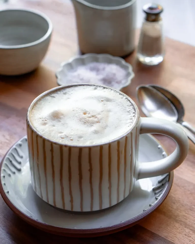 A mug of maple sea salt latte. The mug sits on a small plate. there are small pitchers and a mini bowl of salt behind the mug.