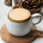 A mug filled with snickerdoodle latte. The mug is on a small round wood board. There are pine cones in a wood bowl behind the latte.