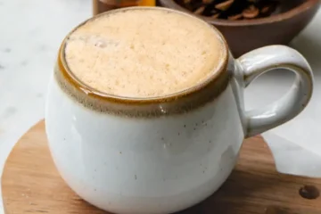 A mug filled with snickerdoodle latte. The mug is on a small round wood board. There are pine cones in a wood bowl behind the latte.