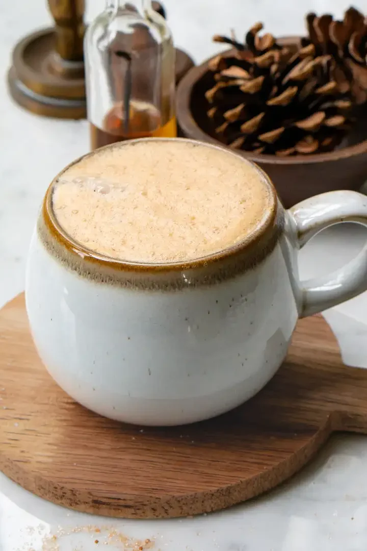 A mug filled with snickerdoodle latte. The mug is on a small round wood board. There are pine cones in a wood bowl behind the latte.