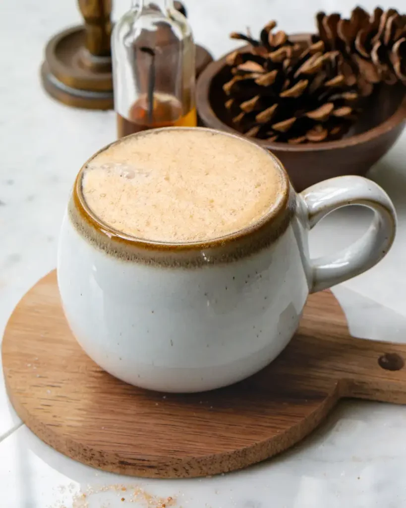 A mug filled with snickerdoodle latte. The mug is on a small round wood board. There are pine cones in a wood bowl behind the latte.