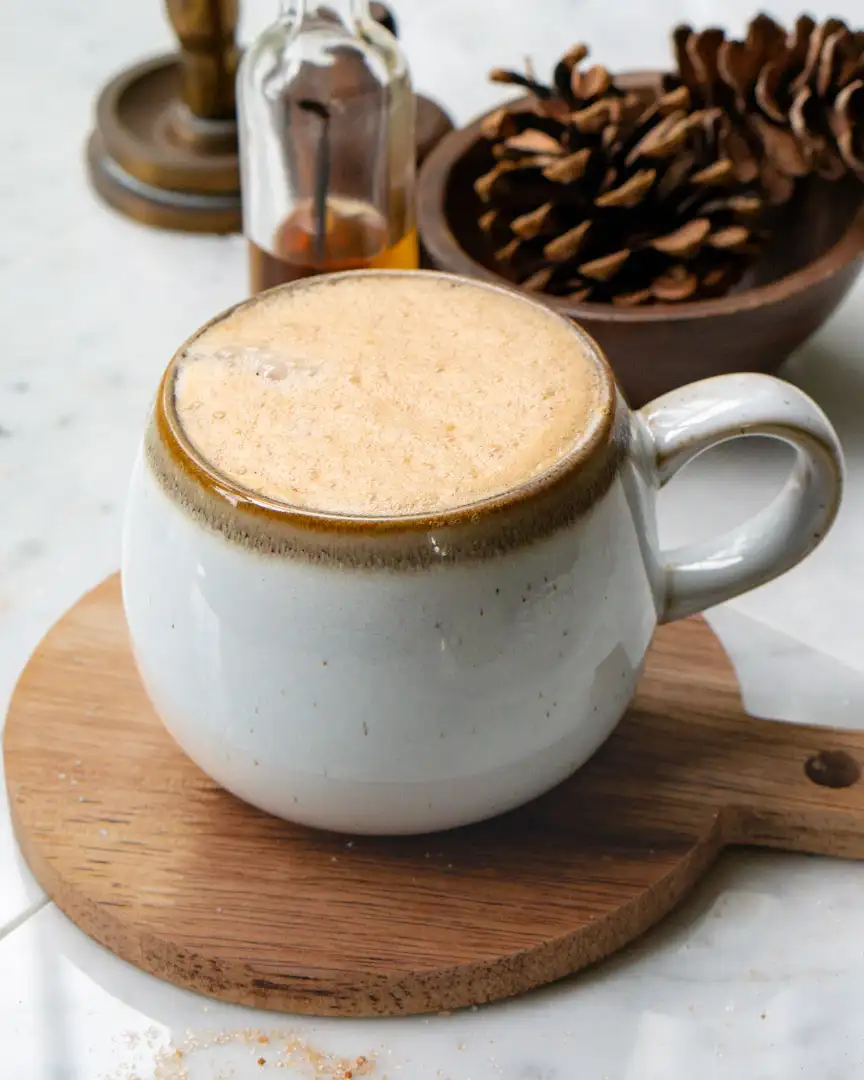 A mug filled with snickerdoodle latte. The mug is on a small round wood board. There are pine cones in a wood bowl behind the latte.