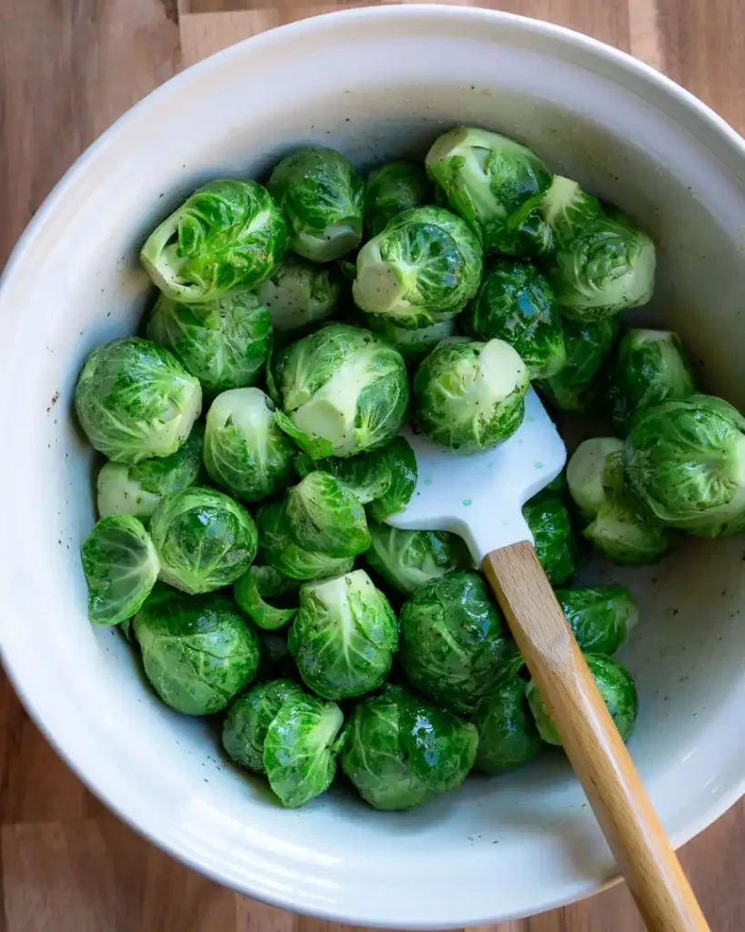 A bowl of raw brussels sprouts that are seasoned. There is spatula in the bowl.