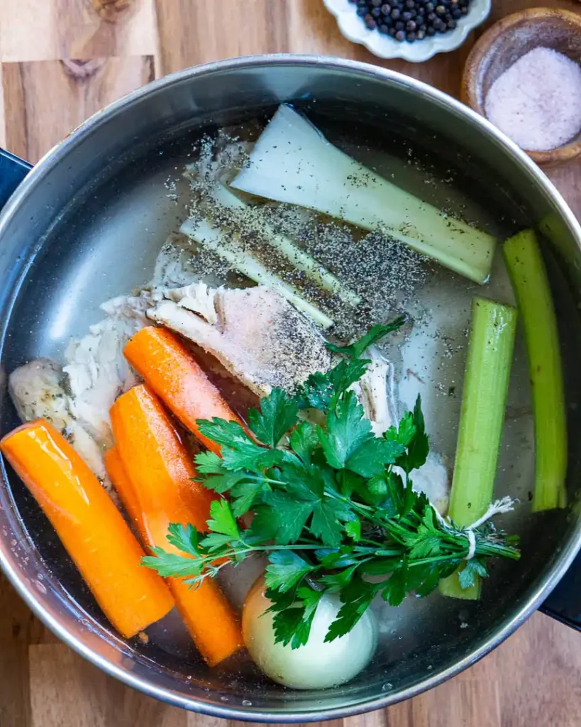 A pot of water with a chicken carcass and raw vegetables in it.