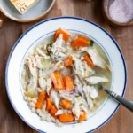 A bowl of chicken soup with a spoon in it. There is a wedge of parmesan cheese, and a small bowls of salt and pepper next to the soup.