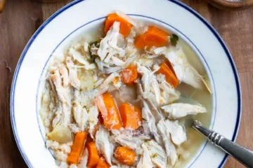 A bowl of chicken soup with a spoon in it. There is a wedge of parmesan cheese, and a small bowls of salt and pepper next to the soup.