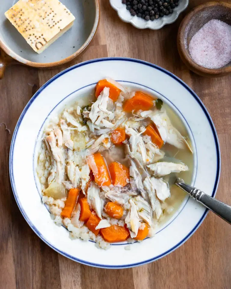 A bowl of chicken soup with a spoon in it. There is a wedge of parmesan cheese, and a small bowls of salt and pepper next to the soup.