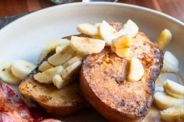 Two slices of french toast on a plate topped with chopped bananas and maple syrup.There is crispy bacon next to it.