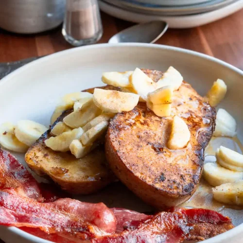 Two slices of french toast on a plate topped with chopped bananas and maple syrup.There is crispy bacon next to it.