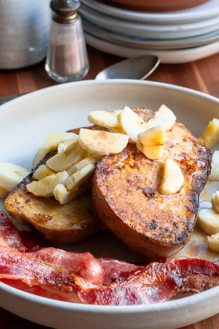 Two slices of french toast on a plate topped with chopped bananas and maple syrup.There is crispy bacon next to it.