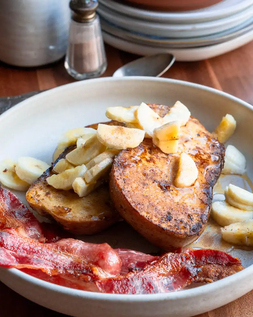 Two slices of french toast on a plate topped with chopped bananas and maple syrup.There is crispy bacon next to it.