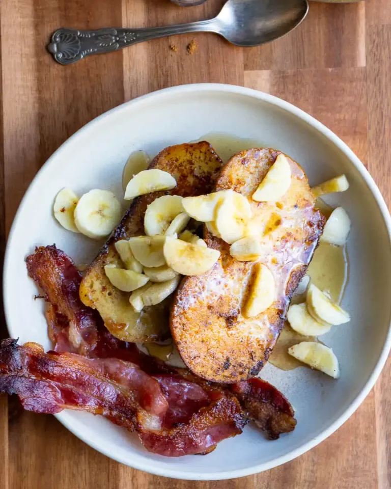 French toast with chopped bananas on top. There is crispy bacon next to it.