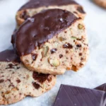 A gluten free shortbread cookie half dipped in chocolate on top of a stack of similar cookies.