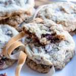 A stack of toffee walnut chocolate chip cookies with the top cookie broken and only a small portion is left. There is a ribbon weaving through the cookies for decoration.