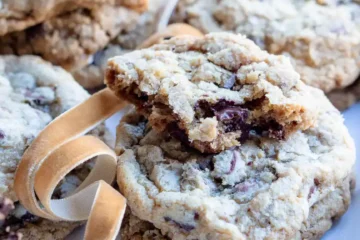 A stack of toffee walnut chocolate chip cookies with the top cookie broken and only a small portion is left. There is a ribbon weaving through the cookies for decoration.