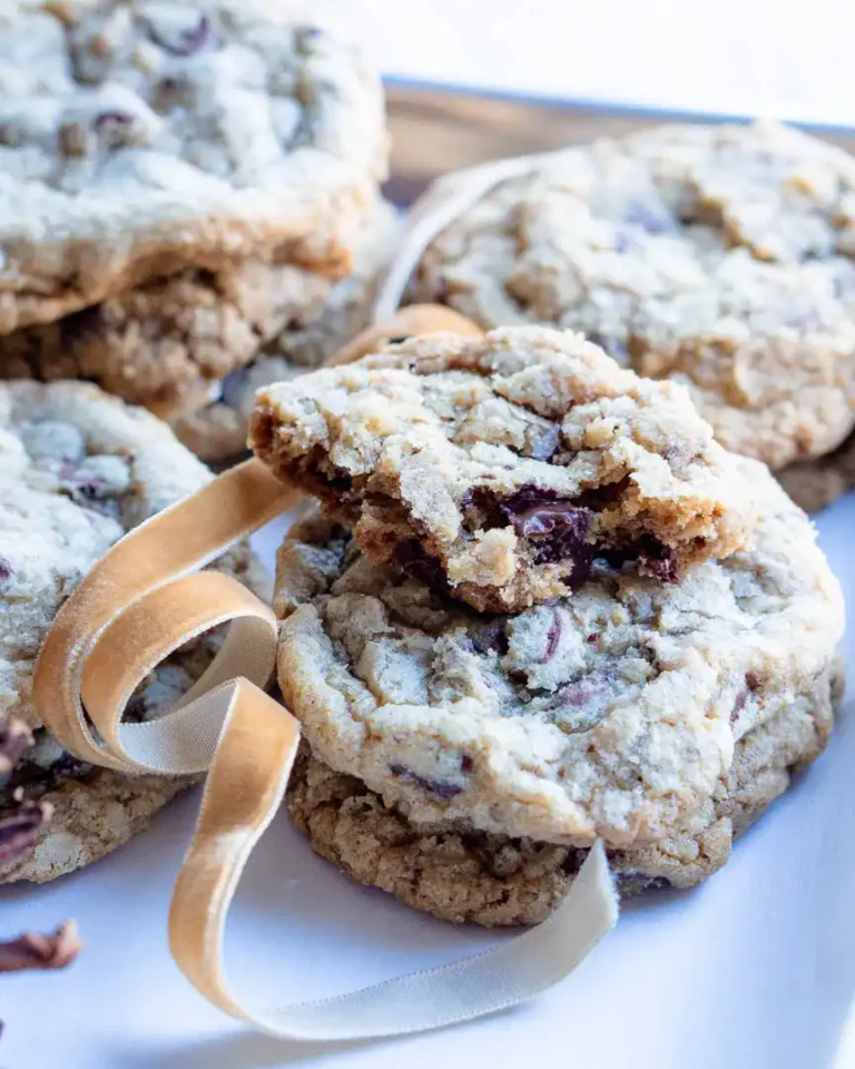 A stack of toffee walnut chocolate chip cookies with the top cookie broken and only a small portion is left. There is a ribbon weaving through the cookies for decoration.