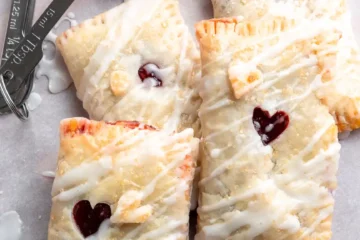 Rectangles of puff pastry filled with cherry filling piled on top of each other. They have a small hole in the middle to let the filling peak out and they have a drizzle of frosting on them.