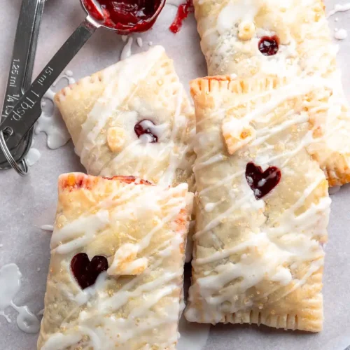 Rectangles of puff pastry filled with cherry filling piled on top of each other. They have a small hole in the middle to let the filling peak out and they have a drizzle of frosting on them.